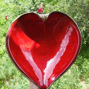 A heart-shaped ceramic bowl with a shiny red glaze on the inside and a brown rim. The exterior is stone-like in brown and beige. The curve of the heart is like a spiral and several red balls descend in a row below it.