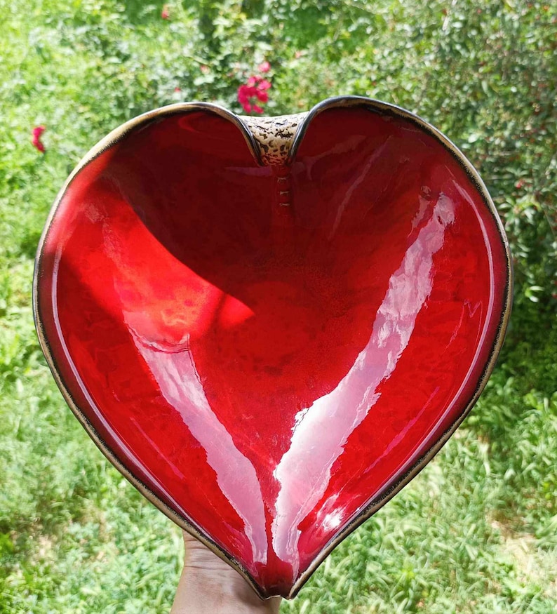 A heart-shaped ceramic bowl with a shiny red glaze on the inside and a brown rim. The exterior is stone-like in brown and beige. The curve of the heart is like a spiral and several red balls descend in a row below it.