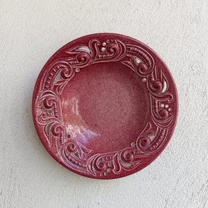May include: A circular, burgundy ceramic plate featuring a detailed, raised floral and scrollwork border. The plate has a textured surface and a slightly glossy finish. The design is symmetrical and adds an elegant touch to the tableware.