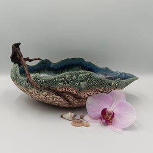 Può includere: Una ciotola in ceramica fatta a mano con un design strutturato e organico. La ciotola presenta un interno blu, un esterno verde e marrone con dettagli di conchiglie e un ramo decorativo in legno. Un'orchidea rosa e conchiglie sono nelle vicinanze.
