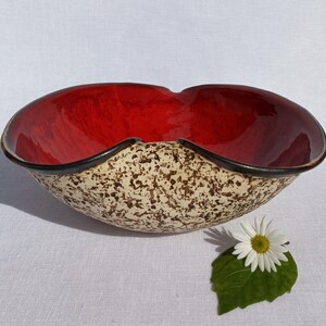 A heart-shaped ceramic bowl with a shiny red glaze on the inside and a brown rim. The exterior is stone-like in brown and beige. The curve of the heart is like a spiral and several red balls descend in a row below it.