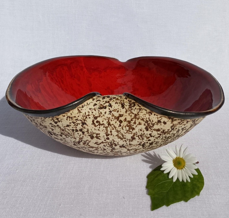 A heart-shaped ceramic bowl with a shiny red glaze on the inside and a brown rim. The exterior is stone-like in brown and beige. The curve of the heart is like a spiral and several red balls descend in a row below it.