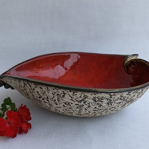 A heart-shaped ceramic bowl with a shiny red glaze on the inside and a brown rim. The exterior is stone-like in brown and beige. The curve of the heart is like a spiral and several red balls descend in a row below it.