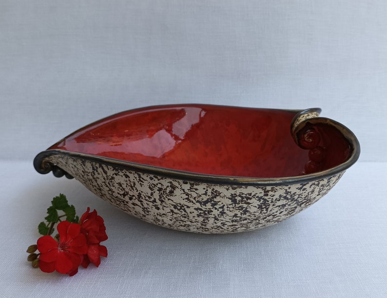 A heart-shaped ceramic bowl with a shiny red glaze on the inside and a brown rim. The exterior is stone-like in brown and beige. The curve of the heart is like a spiral and several red balls descend in a row below it.