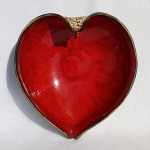 A heart-shaped ceramic bowl with a shiny red glaze on the inside and a brown rim. The exterior is stone-like in brown and beige. The curve of the heart is like a spiral and several red balls descend in a row below it.