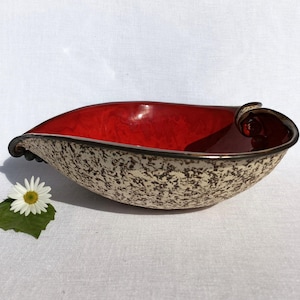 A heart-shaped ceramic bowl with a shiny red glaze on the inside and a brown rim. The exterior is stone-like in brown and beige. The curve of the heart is like a spiral and several red balls descend in a row below it.