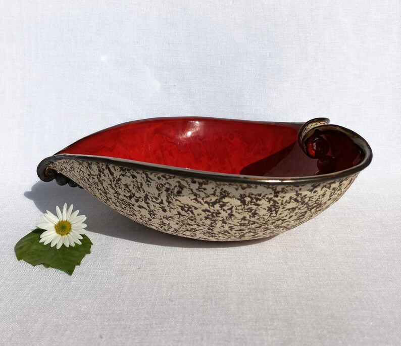 A heart-shaped ceramic bowl with a shiny red glaze on the inside and a brown rim. The exterior is stone-like in brown and beige. The curve of the heart is like a spiral and several red balls descend in a row below it.