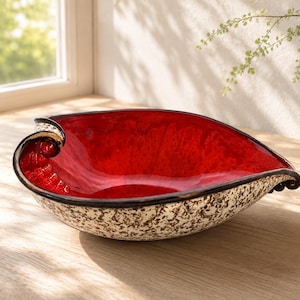 A heart-shaped ceramic bowl with a shiny red glaze on the inside and a brown rim. The exterior is stone-like in brown and beige. The curve of the heart is like a spiral and several red balls descend in a row below it.