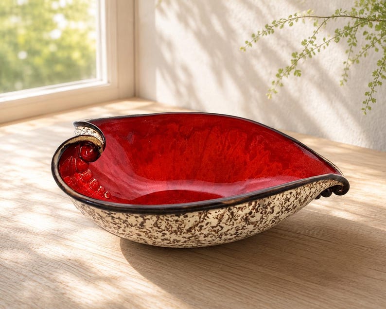 A heart-shaped ceramic bowl with a shiny red glaze on the inside and a brown rim. The exterior is stone-like in brown and beige. The curve of the heart is like a spiral and several red balls descend in a row below it.
