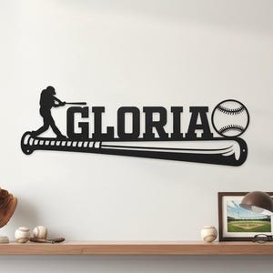 May include: Black metal baseball-themed wall art featuring the name "GLORIA" in bold letters, a baseball, and a batter silhouette. The design is mounted on a white wall, with a wooden shelf below.