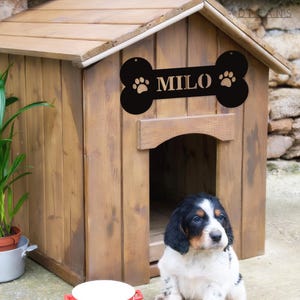 Könnte beinhalten: Ein hölzernes Hundehütte mit einem schwarzen, knochenförmigen Schild mit der Aufschrift "MILO". Das Schild hat an jedem Ende Pfotenabdrücke. Ein kleiner, schwarz-weiß-brauner Welpe sitzt vor dem Hundehütte.