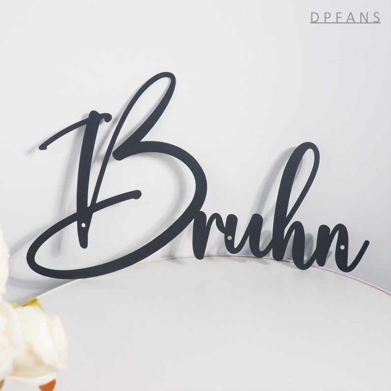 May include: Black metal sign with the name "Bruhn" in a cursive font. The sign is mounted on a white surface, with the letters connected and a decorative style. The word "DPFANS" is in the top right corner.