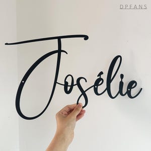 May include: Black metal sign with the name "Jos&eacute;lie" in a cursive font. The sign is held by a hand against a white wall. The sign has two small holes for mounting.