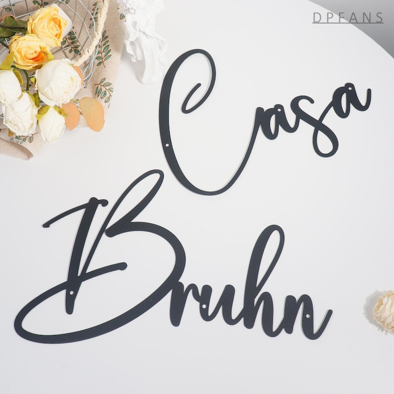 May include: Black metal wall art spelling "Casa Bruhn" in a cursive font. The letters are mounted on a white surface. The image also includes a basket of flowers and a decorative element.