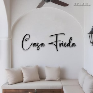 May include: Black metal sign with the words "Casa Frieda" in a cursive font, mounted on a white wall. The sign is above a white cushioned bench with three beige pillows. A ceiling fan is visible in the background.