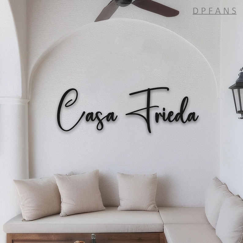 May include: Black metal sign with the words "Casa Frieda" in a cursive font, mounted on a white wall. The sign is above a white cushioned bench with three beige pillows. A ceiling fan is visible in the background.