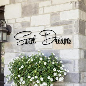 May include: Black metal sign with the words "Sweet Dreams" in a cursive font. The sign is mounted on a stone wall above a planter filled with white flowers and green foliage. A black lantern is mounted on the wall to the left.