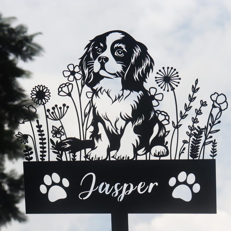 May include: Black metal silhouette of a Cavalier King Charles Spaniel dog sitting in a field of flowers with the name "Jasper" on a sign below.