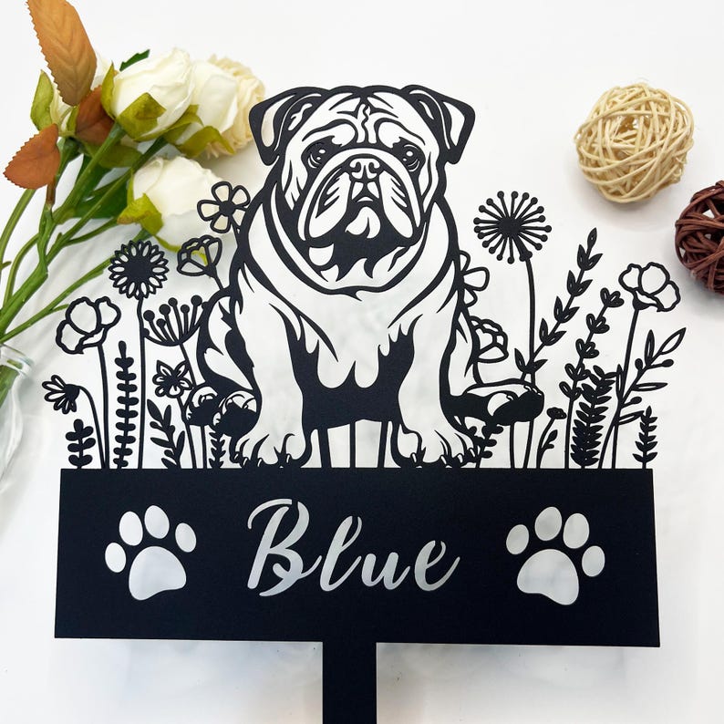 May include: Black metal garden stake featuring a silhouette of a bulldog with flowers and foliage. The stake has a black sign with the name "Blue" and two paw prints.