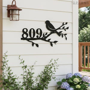 May include: Black metal address sign with the number 809, a bird, and branches. The sign is fixed to a white clapboard wall. A rusty brown outdoor lantern is mounted above the sign.