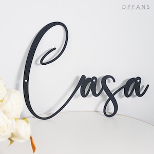 May include: Black metal wall art spelling "Casa" in elegant cursive script. The word is mounted on a white wall, with a small bouquet of white flowers in the lower left corner. The letters have small holes for mounting.