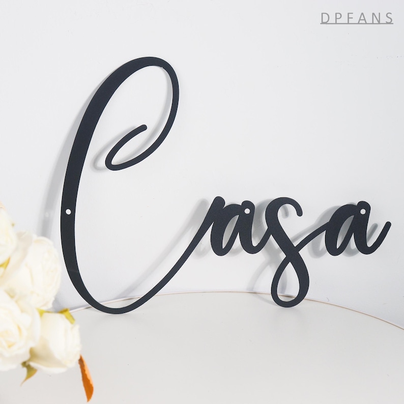 May include: Black metal wall art spelling "Casa" in elegant cursive script. The word is mounted on a white wall, with a small bouquet of white flowers in the lower left corner. The letters have small holes for mounting.