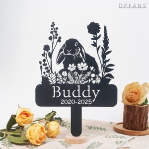 May include: Black silhouette of a rabbit with flowers and the text "Buddy 2020-2025" on a black metal sign with a wooden stake.