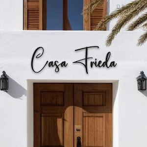 May include: Black cursive metal sign that reads "Casa Frieda" mounted on a white stucco wall above a wooden door. The door is framed by a white trim and flanked by black sconces. A palm frond is visible.