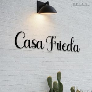 Peut inclure: Panneau métallique noir avec l'inscription "Casa Frieda" en écriture cursive élégante, fixé sur un mur de briques blanches. Un luminaire extérieur noir est au-dessus de l'enseigne, avec un cactus en bas.