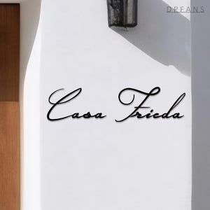 May include: Black cursive text on a white wall reads "Casa Frieda." The text is in a flowing script style. The word "DREANS" is in the upper right corner. A dark metal fixture is above the text.