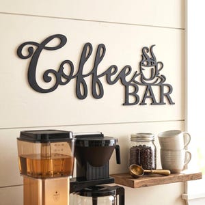 Peut inclure: Panneau décoratif noir "Coffee Bar" avec un motif de tasse à café, monté sur un mur clair. En dessous, une cafetière, un bocal de grains de café, des tasses et une étagère en bois sont visibles.