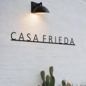 May include: A black metal sign with the text "CASA FRIEDA" on a white brick wall. A black light fixture with a round shade is mounted above the sign.