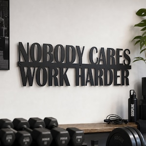 Könnte beinhalten: Schwarzes Metall-Wandbild mit dem Schriftzug "NOBODY CARES WORK HARDER". Das Schild ist an einer weißen Wand befestigt. Unterhalb befinden sich Hanteln, eine Wasserflasche und ein Springseil.