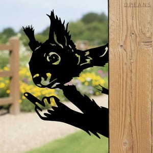 May include: Black metal squirrel silhouette with a raised middle finger, peeking from behind a wooden post. The squirrel's details are cut out, showing the background. The design is suitable for outdoor garden decor.