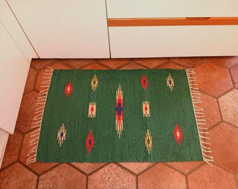 Puede incluir: Una alfombra verde con un patrón geométrico en rojo, azul y amarillo. La alfombra tiene flecos en los bordes.