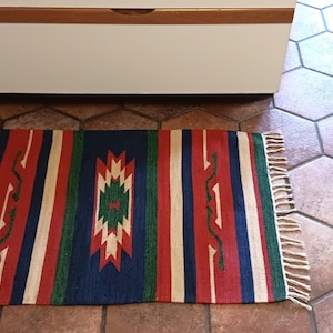 May include: A colorful southwestern style rug with red, blue, green, and white stripes and a geometric pattern in the center. The rug has fringe on the edges.