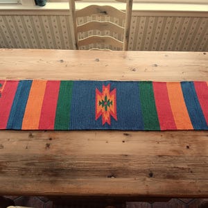 May include: A colorful woven table runner with a geometric pattern in shades of red, orange, green, blue, and yellow. The runner is placed on a wooden table.