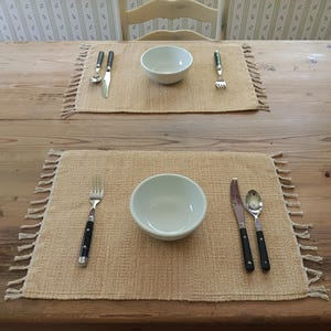 May include: Two place settings with white bowls, forks, knives, and spoons on beige placemats with fringe. The place settings are on a wooden table.