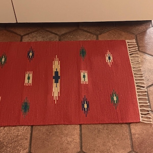 May include: A red rug with a geometric pattern in white, blue, and green. The rug has fringe on the edges.