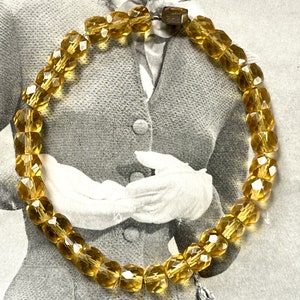 May include: A vintage bracelet with amber-coloured, faceted beads. The beads are strung together, forming a circular shape. A small, silver-toned charm hangs from the bracelet. The clasp is a rectangular shape. The bracelet is displayed against a vintage background.