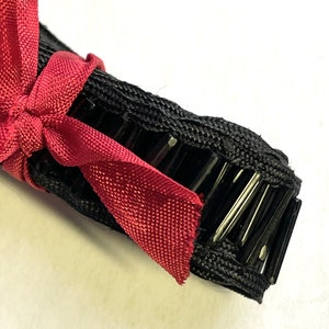 May include: Close-up of a black hair comb with silver metal teeth, tied with a burgundy ribbon. The comb is made of a woven material and is designed for styling hair. The ribbon adds a decorative touch.