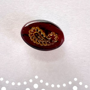 May include: Oval-shaped button with a dark red background and a gold paisley design. The button has a glossy finish and is set against a white background with a decorative edge. The button is a decorative element for clothing or crafts.