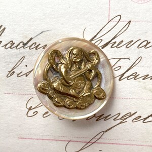 May include: A round button featuring a circular mother-of-pearl base with an iridescent finish. Centered on the button is a raised, metallic gold-toned brass relief depicting a seated figure playing a stringed instrument while resting on clouds.