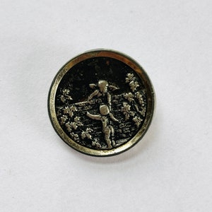 May include: A round, antique silver button with a detailed relief design. The button features two figures in a garden setting, surrounded by foliage. The button has a dark, textured background and a silver border.