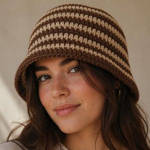 May include: A crochet bucket hat featuring alternating horizontal stripes in dark brown and light beige. The hat has a rounded crown and a short, downward-sloping brim, constructed with a visible textured yarn stitch pattern throughout the design.