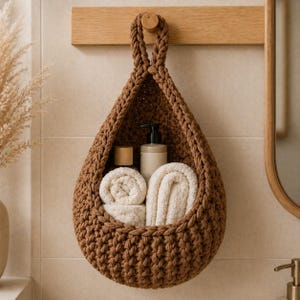 Puede incluir: Una cesta de almacenamiento de ganchillo marrón, en forma de lágrima, colgada de una barra de madera. La cesta contiene toallas blancas enrolladas, una botella con bomba y una botella con tapón de madera.