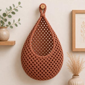 May include: A teardrop-shaped macrame wall hanging basket in a terracotta color. The basket features a diamond-patterned open weave design and is suspended from a single wooden peg by a thick, braided cord loop at the top of the structure.