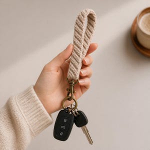 May include: A beige crochet wristlet keychain with a bronze-colored clasp and key ring, holding two car keys. The wristlet has a textured, diagonal pattern. The keys are black and silver.