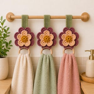 Può includere: Tre portasciugamani a forma di fiore all'uncinetto con anelli di legno e asciugamani abbinati. I fiori sono viola, pesca e gialli, con cinturini verdi. Gli asciugamani sono color crema, verdi e rosa, appesi a una barra di legno.