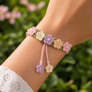 May include: A handmade crochet bracelet featuring a row of colorful flowers in shades of pink, yellow, and purple. The bracelet has a green base and pink ties with dangling flower charms. The bracelet is worn on a wrist.